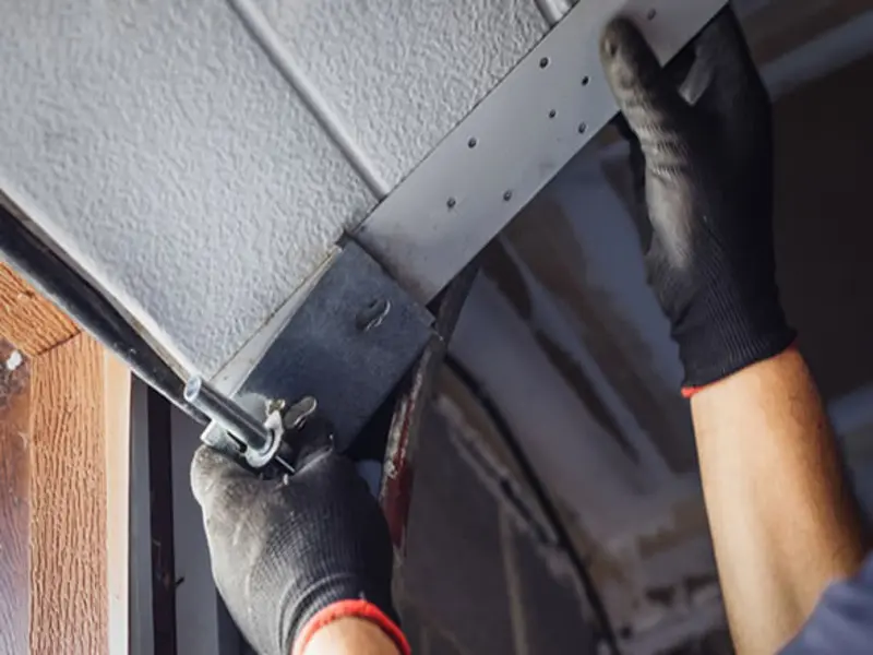 A close-up view of a technician's hands wearing black work gloves while adjusting the brackets and hardware on an overhead garage door panel during a maintenance check.