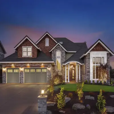 A luxury two-story residential home at twilight featuring a stone facade and a three-car garage setup with premium sage green panel doors. The garage doors include top window inserts and are illuminated by warm architectural accent downlighting under the eaves, presenting high-end residential garage door styling in Odessa, TX.