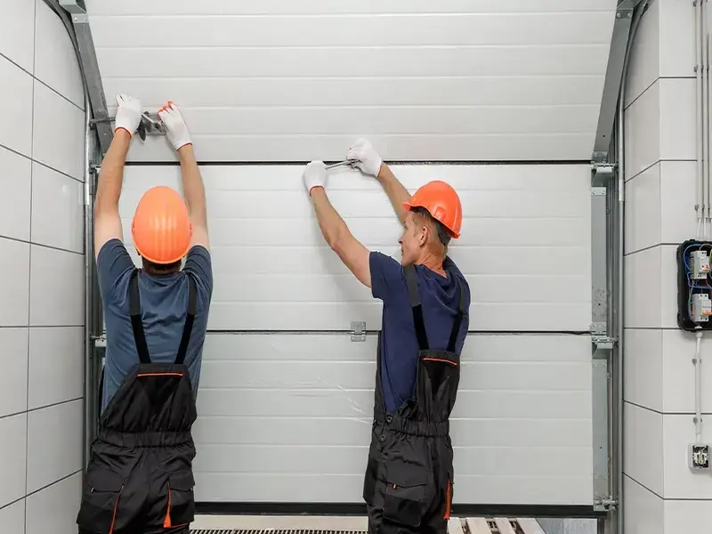 Two garage door technicians wearing orange hard hats and safety gloves work together to repair and align a white sectional garage door on its track.