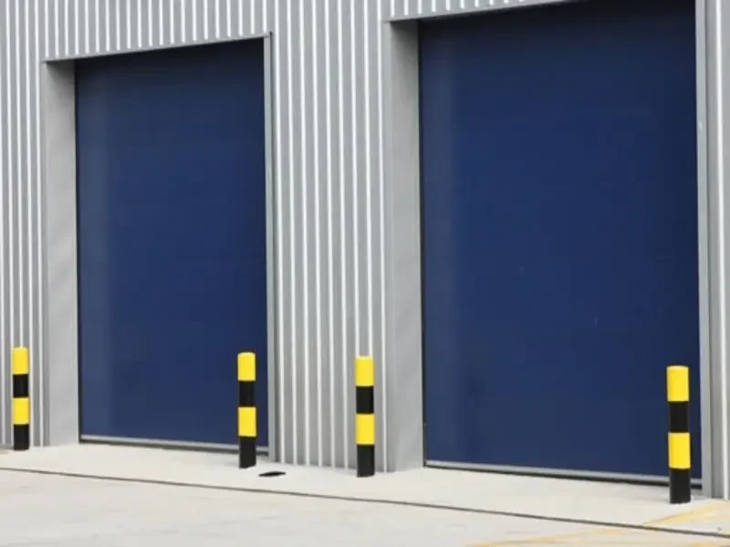 Two large blue commercial roll-up garage doors installed on a corrugated metal warehouse building with yellow and black safety bollards in front.