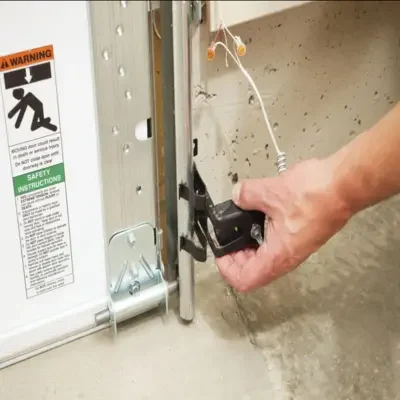 Close-up of a hand adjusting and realigning a black garage door safety sensor with a glowing green indicator light at the base of a vertical metal track during a garage door opener service in Odessa, TX.